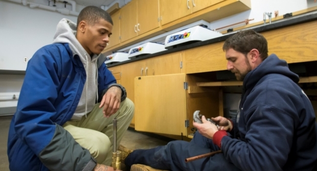 Jamer Jackson (left) shadows university plumber Jason Noel as he repairs a pipe system. | COURTESY Betsy Manning